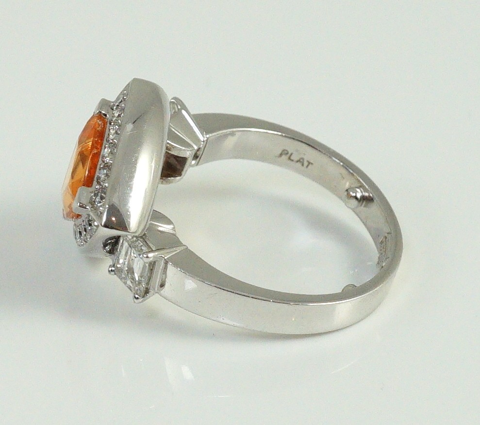 A modern platinum and single stone trilliant cut spessartite garnet ring, with diamond set border and two criss-emerald cut diamond set shoulders, by Musson